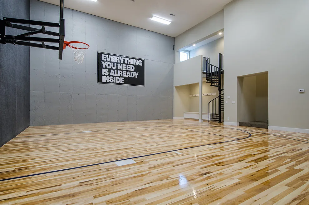 Large basketball gymnasium interior with maple wood flooring, black painted court lines, basketball hoop, and a spiral staircase visible in background